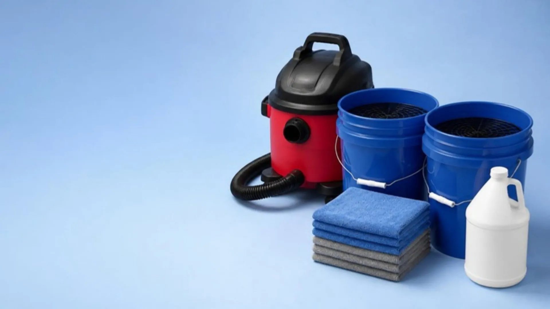A red wet/dry vacuum, two blue wash buckets with grit guards, a stack of microfiber towels and a gallon jug of cleaner