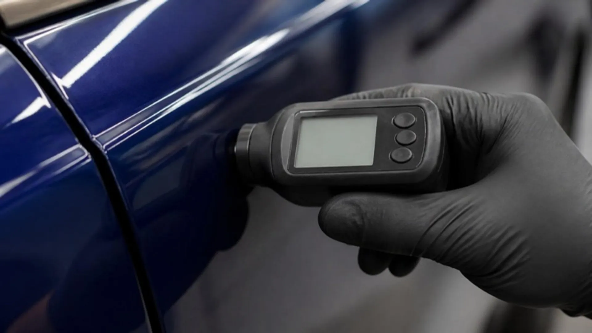 A gloved hand pressing a handheld paint thickness gauge against the door of a dark blue car